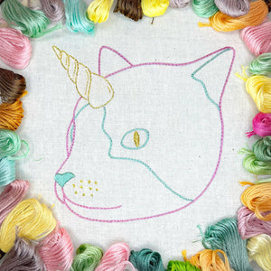 Embroidery hoop with a cat unicorn design surrounded by colorful floss skeins.