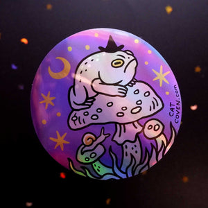 A round sticker featuring a colorful, holographic design of a toad wearing a pointy witch hat sitting on a mushroom under a crescent moon, with the text 'CAT COVEN.COM' printed on the side, set against a dark background with subtle confetti.
