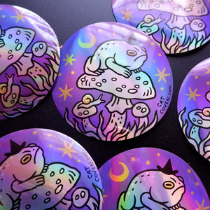 Several round stickers with holographic iridescent finishes, featuring a whimsical illustration of a toad sitting on a mushroom under a crescent moon and stars, displayed on a dark background.