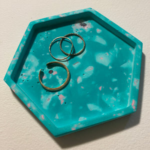 Hexagonal turquoise tray with speckled terrazzo pattern holding three gold rings on a light gray background.