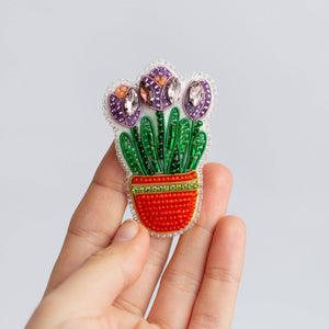 A hand holding a colorful bead embroidered brooch depicting three purple tulips in a red pot with green leaves, set against a plain light gray background.