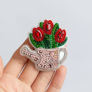 A hand holding a decorative brooch shaped like a watering can with red tulips and green leaves, embellished with pink rhinestones and silver-tone metal, against a light gray background.