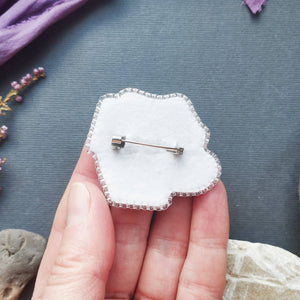 A hand holding a white cloud-shaped brooch with a silver metal border and safety pin back, set against a dark gray background with decorative stones and dried flowers.