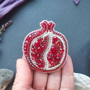 Hand holding a red and silver beaded pomegranate ornament on a textured surface with plants and stones.