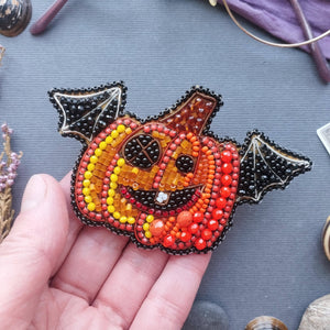 A hand holding a beaded pumpkin-shaped brooch with bat wings. The brooch features a gradient of orange, yellow, and red beads forming a jack-o'-lantern face, with black beaded bat wings and a black outer border, set against a gray textured background with blurred decorative items.