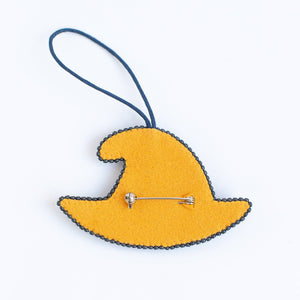 A yellow felt witch hat brooch with a dark blue loop string and a silver safety pin fastener, shown on a plain white background.