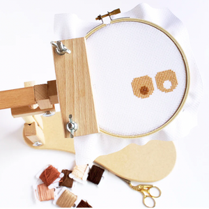 Embroidery hoop with cross-stitched design, wooden frame, scissors, and thread on a white background. The design in the hoop is the "Boob Crew" design in progress.