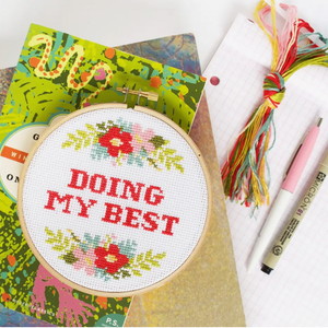 A cross stitch kit with the phrase 'DOING MY BEST' surrounded by a floral pattern, displayed in an embroidery hoop. Embroidery floss and graph paper and some pens are sitting on the right side.