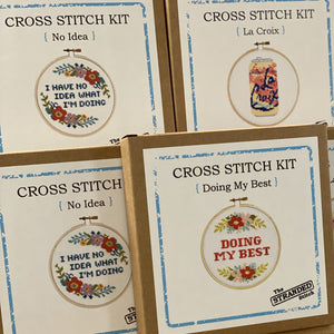 Cross stitch kits with stitched designs on cardboard boxes. They include: I have no idea what I'm doing, La Croix and Doing My Best.