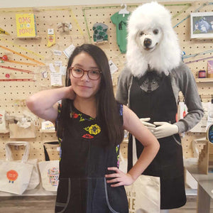 Woman posing with a mannequin wearing a black apron dress in a store setting.
