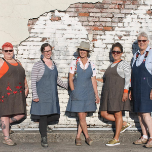 Five women wearing aprons standing in front of a textured wall.