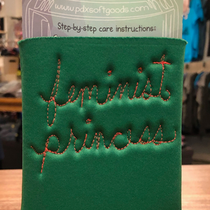 A green fabric can cooler sleeve with 'feminist princess' stitched in orange thread.