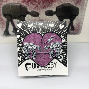 Heart shaped enamel pin called Love AT-AT First Sight with a pink glitter background. Shown on a backing card with hearts.