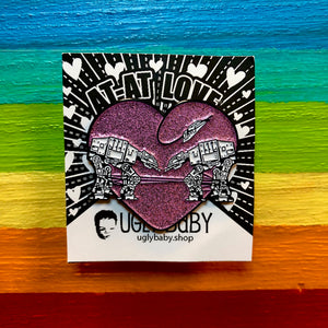 Heart shaped enamel pin called Love AT-AT First Sight with a pink glitter background. Shown against a rainbow background.