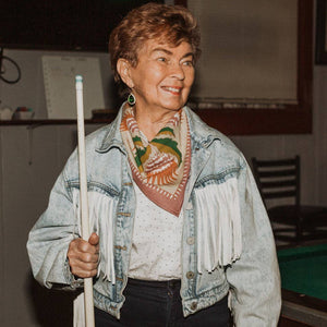 A person wearing a denim jacket and modeling a patterned bandana holding a pool cue.