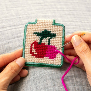 Hand holding a small needlepoint keychain with a red cherry design on a gray fabric background.