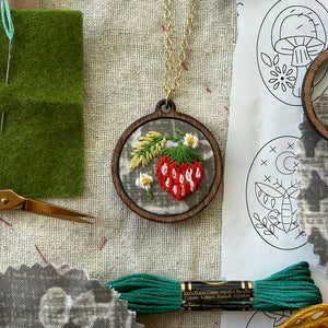 Embroidery hoop with strawberry design on fabric, surrounded by embroidery tools and patterns.