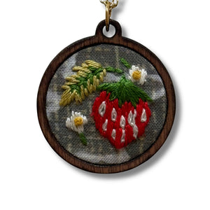 Necklace with embroidered pendant featuring a strawberry and floral design on a wooden background.