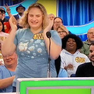 Woman with raised hands on a game show set with audience in the background. She is wearing Ugly Baby's Bubble Cat shirt design.