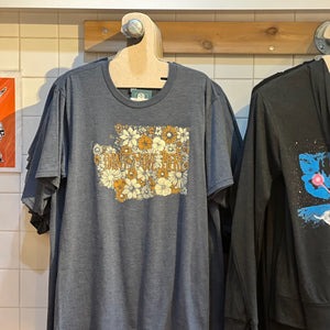 A blue t-shirt with floral design and text that says "Don't Move Here" hanging from a wood display. The floral design is in the shape of Washington state.