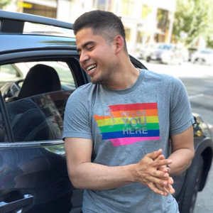 Man wearing a gray t-shirt with a rainbow flag design, standing next to a car.