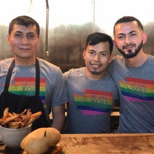 Three men wearing aprons with rainbow designs in a kitchen setting.