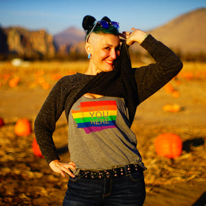 Person wearing a rainbow shirt with 'YOU HERE' text in a pumpkin patch with mountains in the background