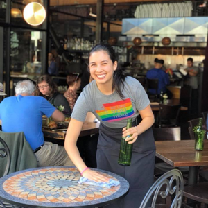 Woman cleaning a table in a restaurant wearing a rainbow tie-dye shirt.