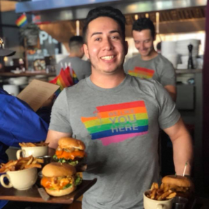 Person wearing a rainbow-themed t-shirt holding burgers and fries in a casual setting