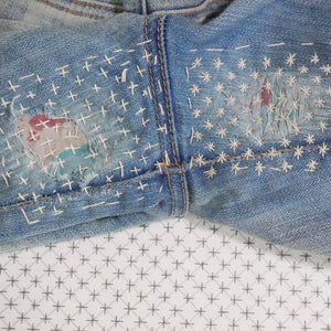 Stitched up example of visible mending on the crotch of a pair of jeans with a stick and stitch pattern in the background.