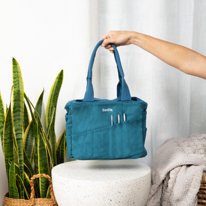 Blue tote bag held by a hand with a plant and blanket in the background