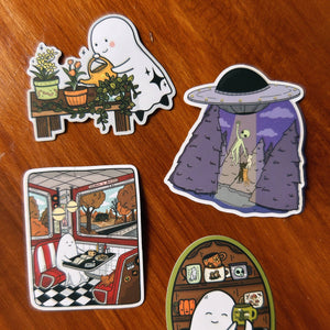 Four cartoon ghost-themed stickers arranged on a wooden tabletop. The stickers show playful scenes including a ghost watering plants, a UFO with an alien, a retro diner interior, and a mug with the words “MUG LOVER.