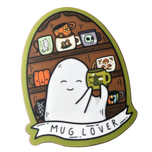 A die-cut sticker featuring a cute white ghost holding a green mug with 'MUG LOVER' written on it, placed on a wooden surface. The sticker has an olive green border and a whimsical coffee shop shelf illustration in the background.