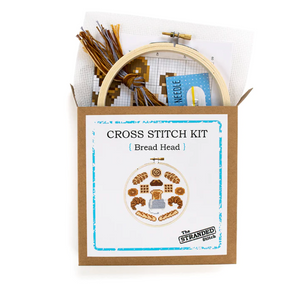The kraft brown box that the Bread Head cross stitch kit by The Stranded Stitch comes in. The supplies that are included in the kit are coming out the top.
