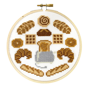 A cross stitch kit with a design that resembles a loaf of bread and various baked goods, displayed in an embroidery hoop.