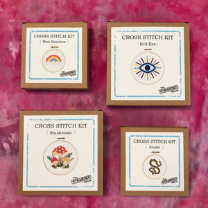 A set of four cross stitch kits laid out on a pink ice dyed tablecloth: Rainbow, Evil Eye, Mushroom & Snake.