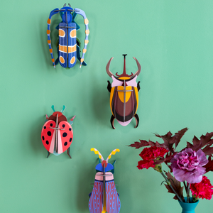 Grouping of bug-themed cardboard wall art sculptures hanging on a bright teal wall with flowers in the foreground.