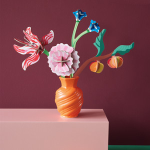 A vase with a variety of paper flowers in it displayed on a pink block against a darker pink wall with nectarines in the foreground.