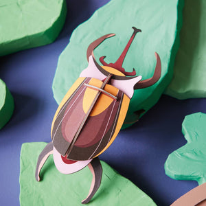 Cardboard sculpture of an elephant beetle on a background of green paper mache leaves.