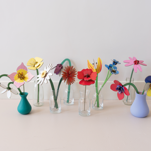 Various colorful flowers made out of cardboard in a variety of vases against a white wall.