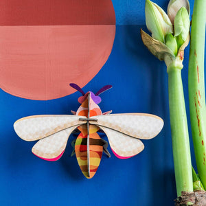 Cardboard sculpture of a honeybee on a blue background next to an amaryllis plant.