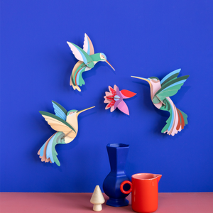Grouping of cardboard hummingbird sculptures on a bright blue wall with multicolored vases in the foreground.