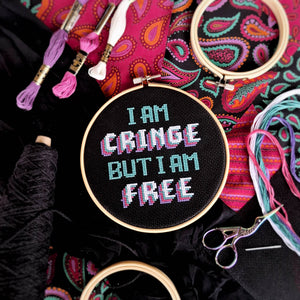 Embroidery hoop with 'I am cringe but I am free' text surrounded by embroidery threads and scissors on a colorful paisley patterned fabric.