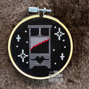 Embroidery hoop with cross-stitched design of a guillotine on a fluffy brown surface, featuring the brand 'The Gloomy Gremlin'.