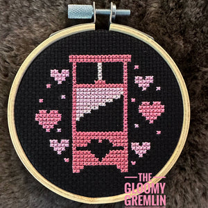 Embroidery hoop with cross-stitched design of a pink guillotine with hearts on a black background, branded 'The Gloomy Gremlin'.