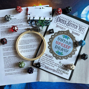 A cross stitch kit laid out on a table with printed instruction sheets, a wooden hoop, thread spools, and polyhedral dice. The main sold item appears to be the cross stitch kit, with the front cover reading 'Cross Stitch Kit' and 'Difficulty: Beginner,' plus 'Untap Upkeep Draw.