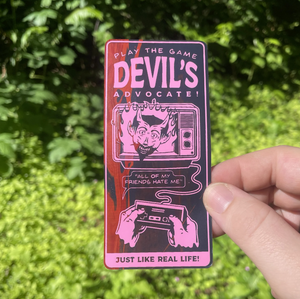 A hand holding a rectangular printed sticker with pink and black graphic art and the text 'DEVIL'S ADVOCATE!' against a blurred green outdoor background.