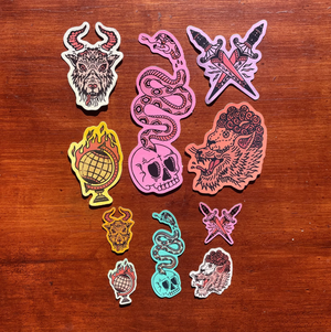 A set of colorful die-cut stickers featuring gothic and edgy illustrations, arranged on a dark brown wooden surface. The designs include a horned goat head, a pink snake with a skull and eye motif, crossed knives with a lightning bolt, a yellow globe with flames, a pink skull, and a lion head.