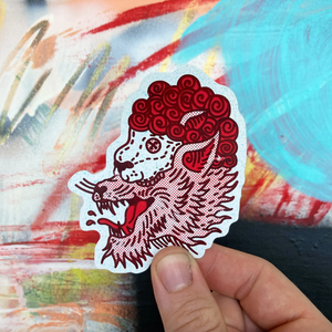 A hand holds a die-cut sticker featuring a stylized red and white wolf in sheep's clothing illustration against a colorful abstract painted background.