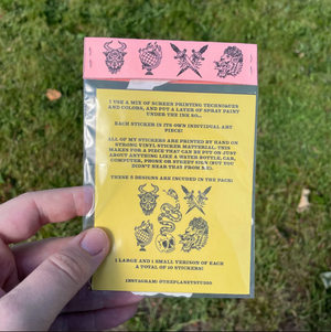 A hand holds a yellow and pink sticker pack card outdoors on grass. The card shows black illustrated designs and printed information about the sticker set, with a visible Instagram handle.